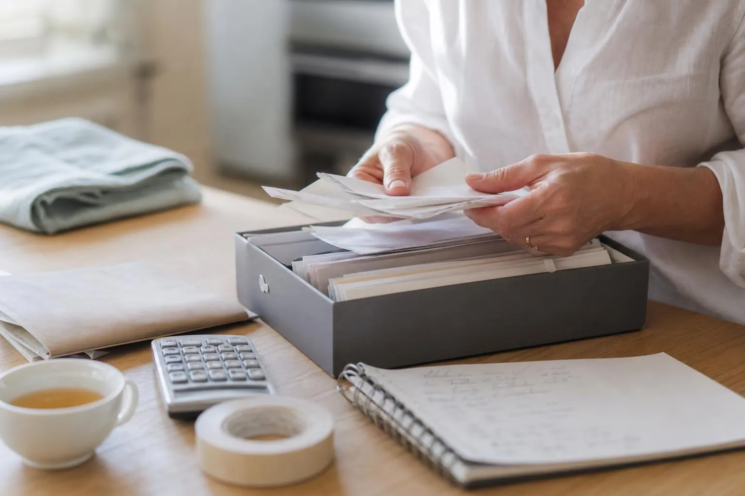 Gros plan sur les mains d’une femme qui trie des tickets et des factures papier au dessus d’une boîte à archives ouverte, sur une table de cuisine claire