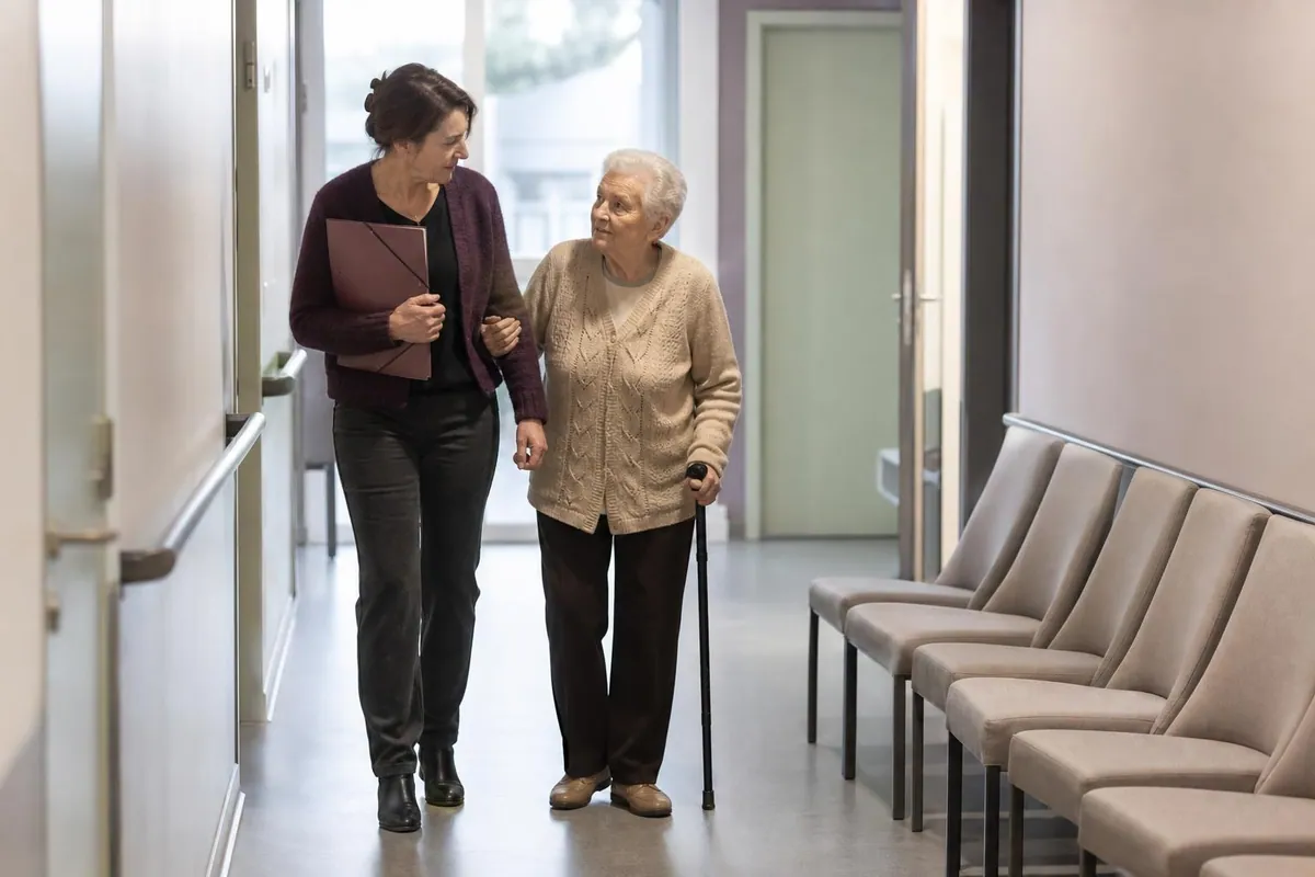 Femme d’une cinquantaine d’années tenant un dossier cartonné et accompagnant sa mère âgée qui marche avec une canne dans le couloir clair d’un centre de santé